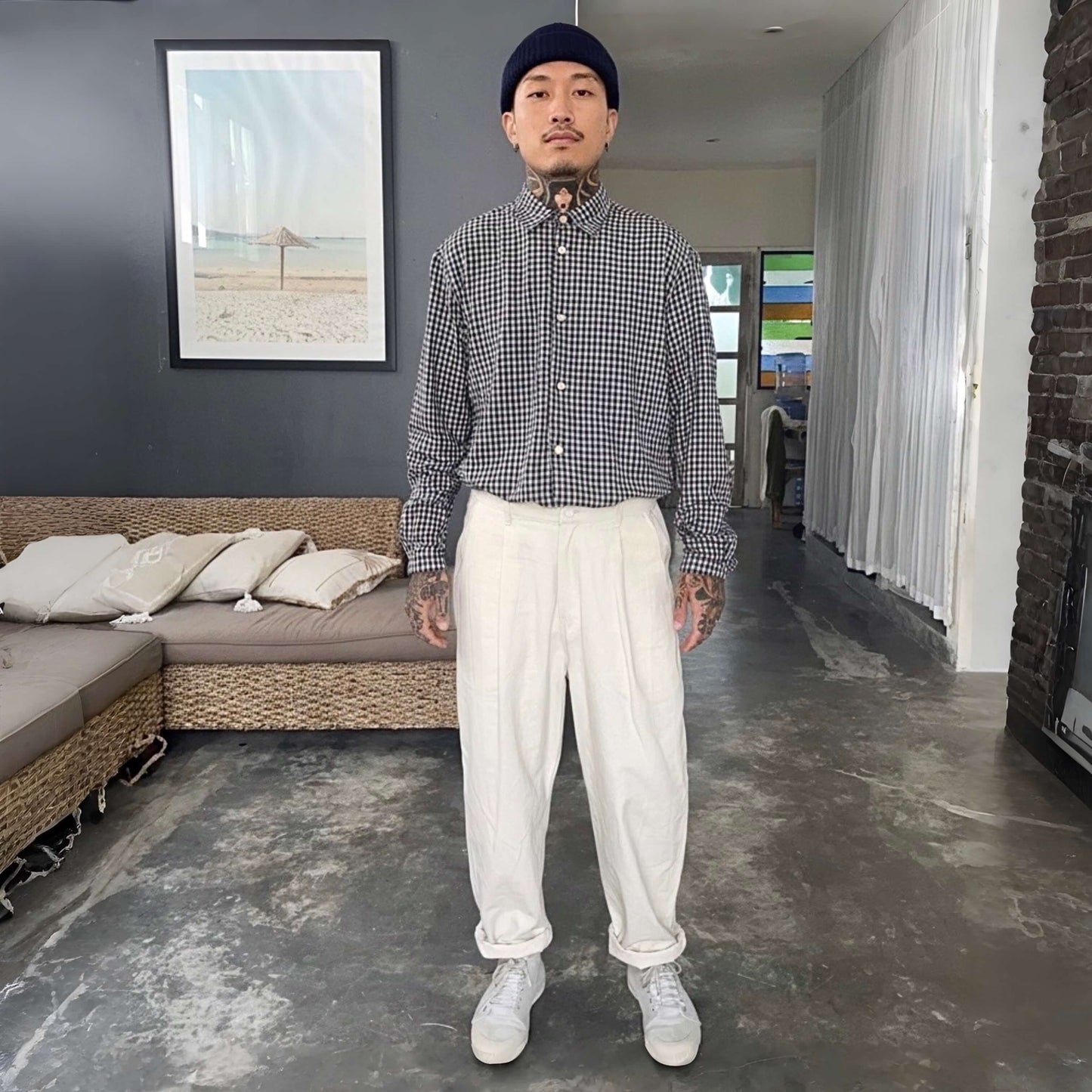 Man standing in a modern living room wearing a checkered shirt, white pants, and a beanie.